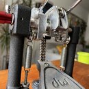 Detailní záběr na mechanismus bicí pedálu s kovovými komponenty stříbrné barvy a černým rámem. Na šlapáku je vyryto logo dw a pod ním částečně vidite…