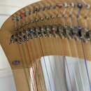 Detailní pohled na horní část harfy z přírodního světlého dřeva. Na krku nástroje jsou viditelné ladicí kolíky a mechanismy s červenými, modrými a bí…