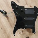 Černý plastový pickguard s třemi integrovanými snímači. Na každém ze snímačů je viditelný nápis emg. Součástí jsou tři černé ovládací knoflíky, přepí…