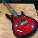 Elektrická kytara s červeným plamenovitým povrchem těla a černým okrajem. Na hmatníku je viditelný nápis "Ibanez". Nástroj má dva humbuckery, most s…