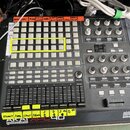 Šedý MIDI kontrolér AKAI professional APL 40 ABLETON CONTROLLER s logem ableton v pravém dolním rohu. Zařízení obsahuje mřížku bílých podsvícených po…
