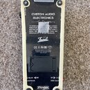 Zadní strana bílého efekt pedálu s černým spodním panelem. Na panelu je nápis CUSTOM AUDIO ELECTRONICS a logo Dual Fasel, dále jsou vidět značky CE,…