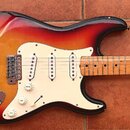 Elektrická kytara s tělem v červeno-žlutém sunburst provedení a bílým pickguardem. Nástroj má tři snímače typu single-coil, jeden přepínač a tři bílé…
