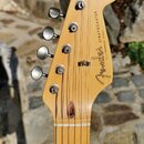 Zadní strana hlavy světle hnědé elektrické kytary s nápisem Fender STRATOCASTER a menším textem WITH SYNCHRONIZED TREMOLO. V horní části je viditelný…