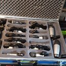 Otevřený hliníkový transportní kufr s šedou akustickou pěnou, v němž je uloženo několik mikrofonů a držáků. Na kufrle je žlutá nálepka s nápisem Mikr…