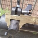 Fotografie zachicuje detail černého kovového svorkovacího mechanismu připevněného na kovovou trubku. K měření průměru je použit zlatavý digitální neb…