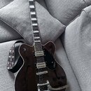 Tmavně hnědá polodublová elektrická kytara s černým pickguardem a bílými lemováním. Na pickguardu je nápis GRETSCH a na hrazdici Bigsby nápis Bigsby.…