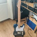 Modrá elektrická kytara s bílým pickguardem a světlou hnedou hmatníkovou deskou. Na hlavě je vidět nápis „Moody Blues“. Kytara stojí na černém stojan…