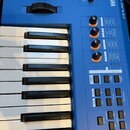 Modrá klávesová novinka s bílými a černými klávesami. Na horní části je nápis YAMAHA a model 64XW, vedle nich se nachází knofík MASTER VOLUME. Pravá…