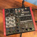 Kompaktní mixovací pult s červeným rámem a černým ovládacím panelem. Na přístroji je nápis DNA PROFESSIONAL CM4-DSP STEREO GRAPHIC EQUALIZER a dále 2…