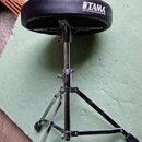Černý bubeníkářský židle s kovovým chromovaným stativem a trojnožkou. Na sedáku je bílý nápis TAMA a pod ním text THE STRONGEST NAME IN DRUMS. Povrch…