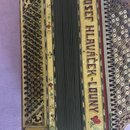 Žlutý akordeon s květinovými ornamenty. Na pravém boku je svislý nápis JOSEF HLAVÁČEK-LOUNY. Nástroj vykazuje známky opotřebení v podobě drobných škr…
