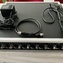Na černé ploše kufríku s hliníkovým rámem leží černý napájecí adaptér, černý kabel s konektory XLR na obou koncích a černý napájecí kabel. V dolní čá…