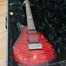 Elektrická kytara s červeným plamenovitým povrchem těla, uložená v černém polstrovaném kufru. Nástroj má dva humbuckery, fixační most a šest ladicích…