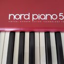 Detail červeného digitálního piana s bílými a černými klávesami. Na horní části je bílý nápis nord piano 5 a pod ním text VIRTUAL HAMMER ACTION TECHN…