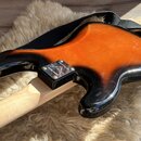 Zadní strana elektrické kytary s černým lakem a oranžovým středem v provedení sunburst. Na kytaru je nasazen černý popruh a vidět je světlá dřevěná h…