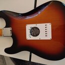 Zadní strana elektrické kytary s barevným přechodem od žluté přes hnědou k černé (sunburst). Na zadní straně těla je bílý kryt elektroniky s nápisem…