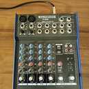Mixážní pult s modrým šasi a stříbrným předním panelem. Na horní části je nápis PRONOMIC M-602 PREMIUM 6-INPUT 2-BUS MIXER. Zařízení obsahuje různé v…