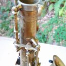 Detailní záběr na horní část saxofonu v matném mosazném provedení. Nástroj vykazuje výrazné opotřebení povrchu, oxidaci a nečistoty. Na fotce nejsou…
