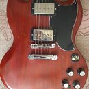 Elektrická kytara s červeným dřevěným tělem a černým plechtrzem. Nástroj má dva humbuckery s kovovými kryty, chromovaný most a stopku. Na ovládacím p…