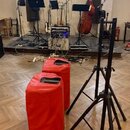 Fotografie zachicuje hudební vybavení v interiéru. V popředí jsou dva červené cylindrické kryty z textilie s úchyty a černý stativ. V pozadí jsou vid…