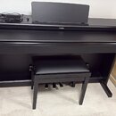 Černý digitální piano v černém nábytkovém stojanu s přívodním kabelem a doprovodnou černou stolečkou. Na přední straně je umístěno logo YAMAHA. Nástr…