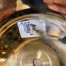 Detail spodní strany kovového činely zlatavé barvy s viditelnými koncentrickými rýhami. Na povrchu je nalepená bílá nálepka s čárcovým kódem a textem…