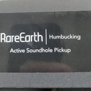 Fotografie detailu černé krabice s bílým textem. Je na ní uveden nápis RareEarth | Humbucking a pod ním Active Soundhole Pickup. Povrch krabice je le…