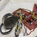 Fotografie zobrazuje elektronickou součástku v kovovém pouzdře připojenou k červené desce s dalšími komponenty a barevným zapojováním kabelů. Na kovo…