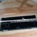 Stříbrná flétna položená v černém otevřeném pouzdře na dřevěném stole. Nástroj má kovový povrch s mechanismem klapek a chybí na něm viditelný nápis z…