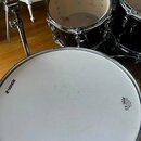 Fotografie zobrazuje část černé bicí sady. Na bílém bubnovém koži v popředí je vidět logo SONOR a nápis UN DRUM HEAD MADE IN CHINA. V pozadí jsou vid…
