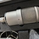 Stříbrný kondenzátorový mikrofon v černé polstrované přepravce. Na těle je viditelný nápis SUPPLY N 149, SER. NO (za kterým následuje nečitelný kód)…