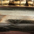 Detail zlatavého kovového povrchu hudebního nástroje. Na povrchu je vidět rytý ornament a několik drobných škrábanců. Žádné značky ani nápisy nejsou…