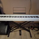 Bílá digitální klaviatura umístěná na černém X-stojanu. Na ovládacím panelu je nápis Casio a řada tlačítek s nečitelnými popisky. Vedle klaviatury st…