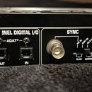 Detail zadní strany černého elektronického zařízení s nápisy. V sekci 8-CH. UNEL DIGITAL I/O jsou dva optické konektory označené jako OUT a IN s nápi…
