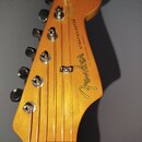 Fotografie zachycuje hlavu elektrické kytary z přírodního dřeva s viditnou sletou. Na povrchu je černý nápis Fender a STRATOCASTER, doplněný menšími…