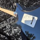 Černá elektrická kytara s tvarem typu Stratocaster v otevřeném černém kufru. Na zadní straně těla je kovový kryt s viditelnými škrábanci a opotřebení…