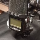 Černý digitální záznamový přístroj značky ZOOM s kovovou mřížkou mikrofonů v horní části. Zařízení má malý LCD displej a je připevněno na černý stati…