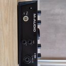 Černý zvukový interface značky M-AUDIO. Na přední straně jsou vidět čtyři otočné knoflíky, jeden velký centrální regulátor a dva výstupní konektory M…