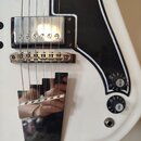 Detail bílé elektrické kytary s černým plechtrádem. Nástroj má chromovaný hmyzub, kobylku a jeden viditelný humbucker s kovovým krytem. Na ovládacích…