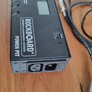Černý kovový napájecí zdroj s nápisem ROCKBOARD BY WARWICK POWER PIT. Na horní straně je umístěn malý displej a čtyři kontrolky s popisky. Na zadní s…