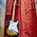 Elektrická kytara s tónem sunburst a bílým pickguardem v otevřeném kufru s červenou plišovou výstelkou. Na hlavě hředu je viditelné logo Fender. Kyta…