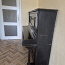 Černý vzpravený piano v matném provedení. Povrch je viditelně opotřebovaný s četnými škrábancemi a odřapáním, zejména na hranách a bočnici. Na nástro…