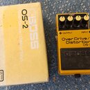 Žlutý kytarový efekt BOSS OS-2 Over Drive/Distortion v originální žluté krabici. Na pedálu jsou čtyři ovládací knoflíky, červená kontrolka CHECK a ná…