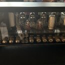 Černý kytarový zesilovač s průhledným předním panelem, za kterým jsou vidět elektronické lampy. Na skle je bílý nápis Hughes & Kettner a vpravo menší…