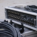 Černý stagebox v hliníkovém transportním kufru. Na přední straně je nápis MIDAS DL16, řada vstupních konektorů a ovládací prvky s popisky HEAD AMP (H…
