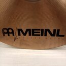 Detail činely z mosazného kovu s koncentrickými drážkami. Na povrchu je černý nápis MEINL a vedle něj logo složených dvou písmen M. Pod hlavním nápis…