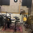 Fotografie zachycuje hudební studio s elektronickou a akustickou bicí sadou. Elektronická sada je černé barvy, na jednom z činelů je vidět nápis Ista…