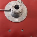 Detail červeného polstrovaného sedáka s kovovým mechanismem uprostřed. Na kovovém diskovém podkladu je viditelná bílá nálepka s číslem 2101. Okraje s…