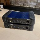 Dvě pasivní DI boxy značky PROEL, model DB1+P PASSIVE D.I.BOX. Horní kus má modré kovové tělo s žebrovaným povrchem a černé plastové kryty na koncích…